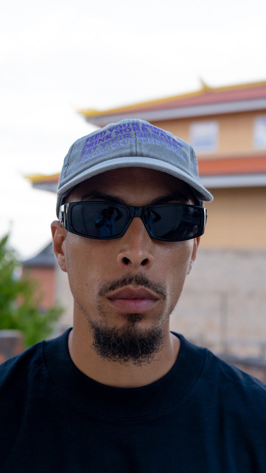 A man wearing a gray baseball cap with purple text, black sunglasses, and a black t-shirt, photographed outdoors with a blurred building in the background. MINOR FORMULA